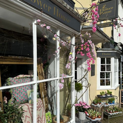 The shop window of the Flower House, Shipston with large window panes, trailing pink flowers and a hanging shop sign saying Flower House, Florist, Gifts and Cards